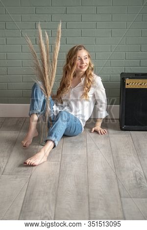 Young Beautiful Woman With Bare Feet Sitting On The Floor Next To Subwoofer And Glass Vase With Dry 