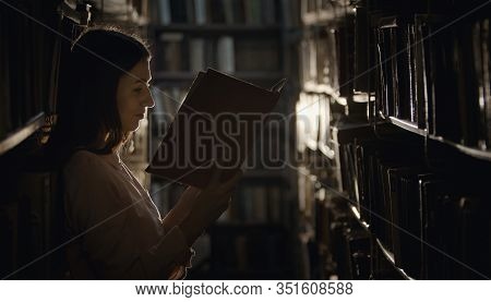 Young Woman Rapt In Reading In Dark Library Standing Among Rows Of Shelves With Books