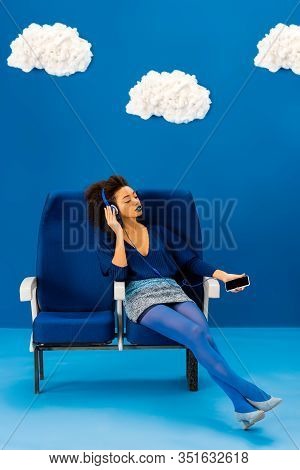 African American Sitting On Seat, Listening To Music And Holding Smartphone On Blue Background With 