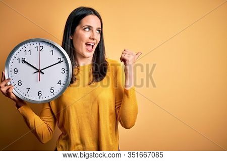 Young woman with blue eyes doing countdown holding big clock over yellow background pointing and showing with thumb up to the side with happy face smiling