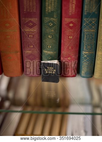 A Small Holy Book Of The Bible Stands On A Shelf Near Large Multi-colored Books.