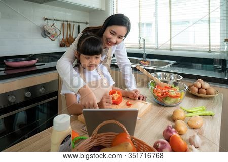 Portrait Of Beautiful Asian Young Woman And Her Daughter Cooking Salad For Lunch Using Online Intern