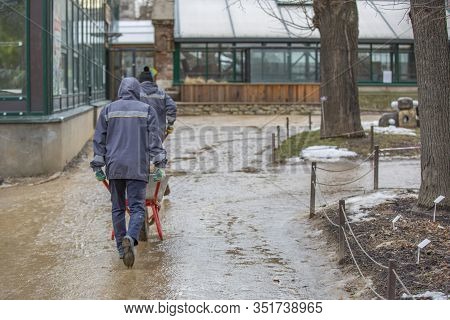 Two Adult Workers In Uniform With Trolleys In A City Park