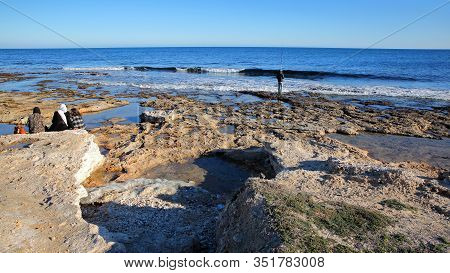 Mahdia, Tunisia - December 26, 2019: A Fisherman And Local Women On The Seashore At The Top End Of T