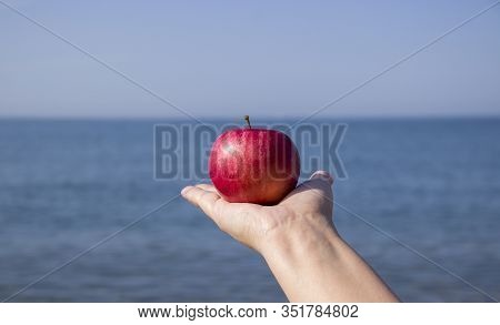 Red Ripe Apple In Hand Against The Sea. Ecotourism And Consumption Of Natural And Organic Fruits.