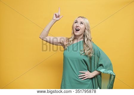 Happy Blond European Woman In Green Dress Thinking Abour Creative Idea, Pointing Up Over Yellow Back