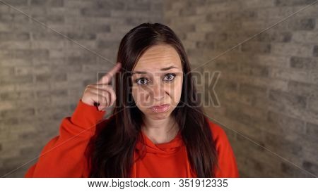 A Beautiful Young Woman In A Red Hoodie Twists At Her Temple On A Gray Brick Background.