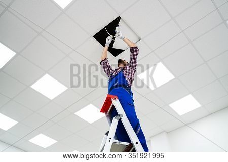 Full Length Portrait Of Electrician On Stepladder Installs Lighting To The Ceiling In Office