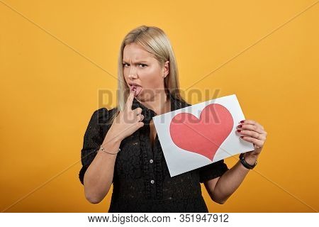 Young Blonde Girl In Black Dress On Yellow Background Angry Woman Holds Piece Of Paper With Red Hear