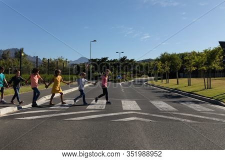 Side view of a diverse group of elementary school pupils crossing an empty road together, half way across a pedestrian crossing, holding hands on a sunny day