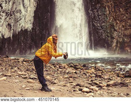 A Woman Stands On The Bank Of A River Against The Backdrop Of A Stormy Waterfall, Filling Her Hat Wi