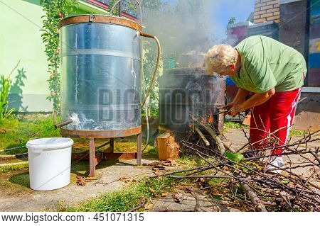 Grandma Throws Dry Branches Into The Firebox Of A Homemade Distillery Making Moonshine Schnapps, Alc