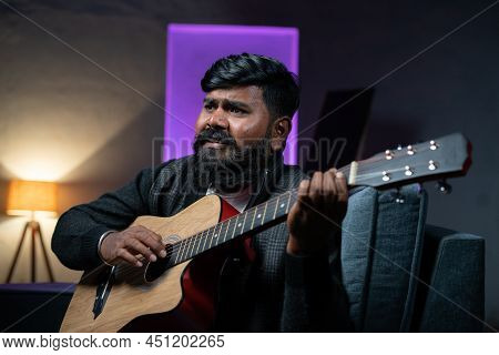 Young Beardman Playing Guitar While Sitting On Sofa At Home - Concept Of Musician, Artist And Hobbie