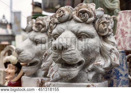 A pair of antique decorative lion head plant pots, with roses forming the mane. Other antiques and bric-a-brac can be seen in the background of a market stall.