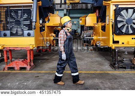 Young female technician in overalls, harhat and gloves moving along large industrial machines standing in rows in warehouse