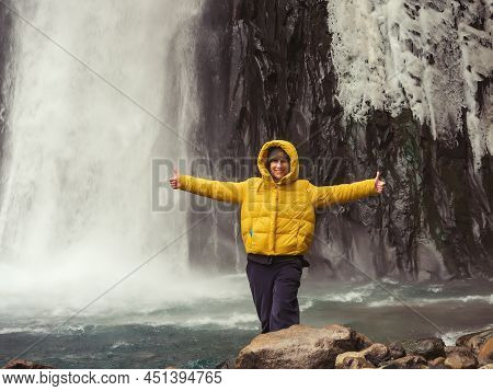 Smiling Woman Showing Thumbs Up Against The Background Of An Icy Waterfall In A Rocky Area