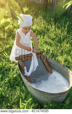 Vertical Photo Of A Child Laundry White Clothes In An Old Basin On A Washboard. Girl Wrings Out Unde