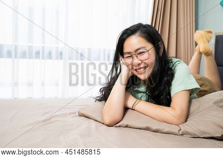 Beautiful Glasses Asian Woman Is Lying On Her Stomach And Looks At Camera On Her Bed  In The Bedroom