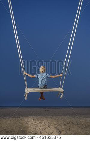 Child Sits On Rope Swing And Looks At The Sea And Majic Dark Blue Sky. Boy Swinging During Dusk. Bac