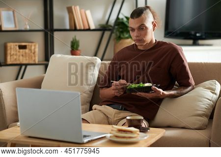 Young Man Eating Salad And Watching Show Episode Or Presentaion On Laptop When Resting On Sofa At Ho
