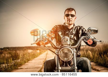 Biker man wearing a leather jacket and sunglasses sitting on his motorcycle looking at the sunset.