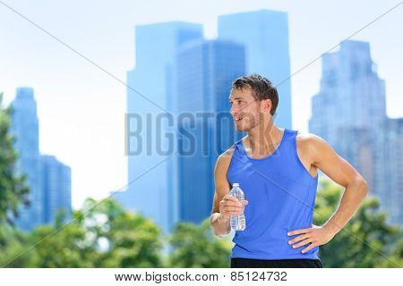 Sport man drinking water bottle in New York City. Male runner sweaty and thirsty after run in Central Park, NYC, Manhattan, with urban buildings skyline in the background.