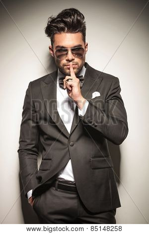 Young business man leaning on a wall showing the silent gesture while holding one hand in his pocket.