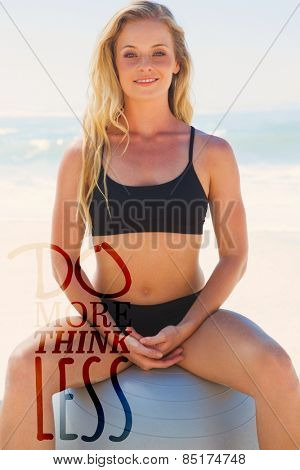 Fit blonde sitting on exercise ball at the beach smiling at camera against do more think less