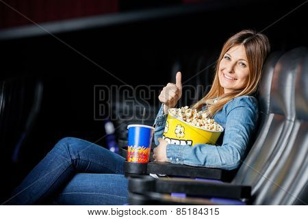 Side view portrait of happy woman showing thumbsup while holding snacks at cinema theater