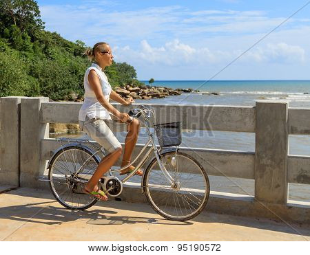 Young Woman Riding Bicycle Across River Bridge Next To Tropical Park