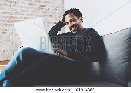 Cheerful bearded African man working on laptop while sitting sofa at his modern home office.Concept of young people enjoying mobile devices.Brick wall blurred background