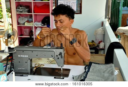 Hua Hin Thailand - January 1 2010: Barechested tailor using a Singer Sewing machine threads a needle at his roadside shop