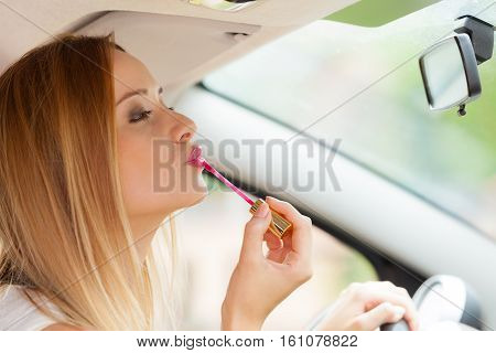 Distracted driver. Young attractive woman looking in rear view mirror painting her lips doing applying make up while driving the car.