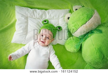 little boy in knitted animal hat lying on soft green blanket with frog softtoy. Cute 5 month old baby on the pillow in bed