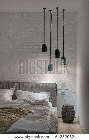 Fancy bedroom in a modern style with white brick wall and a parquet on the floor. There is a bed with a white pillows and a blanket and a brown plaid, lampshade, glowing hanging lamps. Vertical.