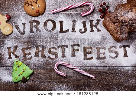 high-angle shot of a wooden table sprinkled with icing sugar or flour where you can read the text vrolijk kerstfeest, merry christmas in dutch, some candy canes and some christmas cookies