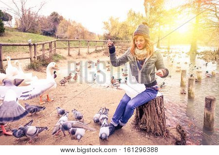 Young woman feeding ducks at the lake park before sunset - Girl in the winter time having fun with with animals by the river - Concept of outdoor life in the cold season - Backlit and sun halo filter