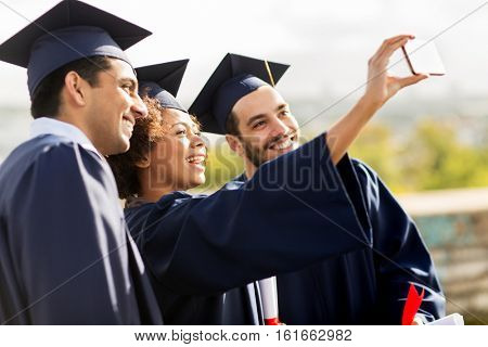 education, graduation, technology and people concept - group of happy international students in mortar boards and bachelor gowns with diplomas taking selfie by smartphone outdoors