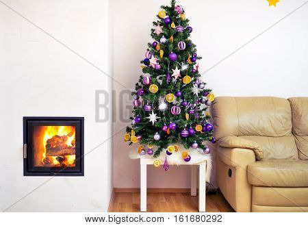 Christmas tree in modern luxury house apartment with fireplace. Sofa next to home decorated Christmas tree with hot fire and flames burning in the fireplace.