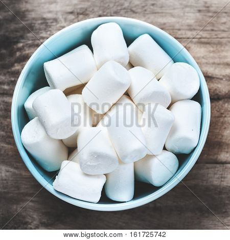 Fluffy white marshmallow in blue round bowl on old wooden table with copy space close up