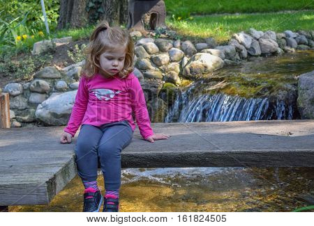 Cute little girl sitting on the old wooden bridge looking at the water in japaneze garden on the beautiful sunny day