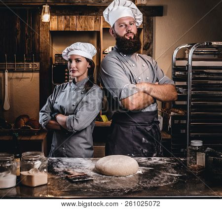 Chef With His Assistant In Cook Uniform Posing With Crossed Arms Near Table With Ready Dough In The 