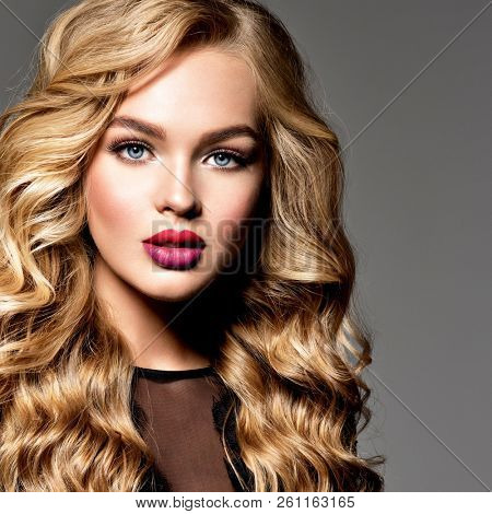 Blond woman with long curly beautiful hair. Makeup. Fashion make-up. Fashionable girl dressed in black dress. Closeup portrait. Gorgeous face of an attractive fashion model. Bright red lips.