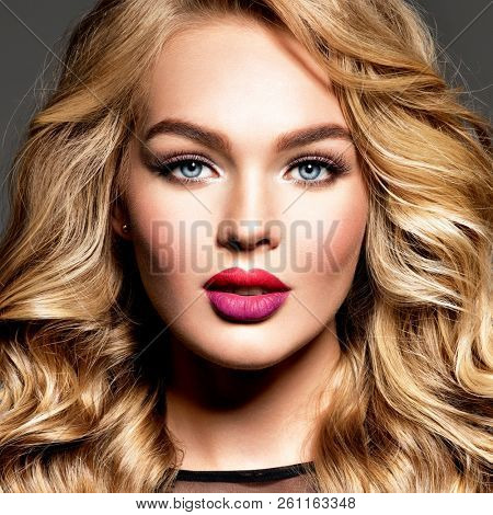 Blond woman with long curly beautiful hair. Makeup. Fashion make-up. Fashionable girl dressed in black dress. Closeup portrait. Gorgeous face of an attractive fashion model. Bright red lips.