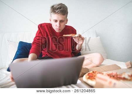 Attractive Young Man With A Piece Of Pizza In His Hands Sits On A Bed With A Box Of Pizza, Watching 