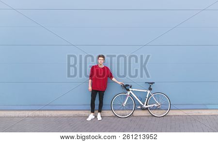 Young Man In Stylish Clothes Is Standing With A White Bicycle On The Background Of A Blue Wall And L