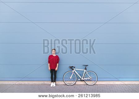 Minimalist Portrait Of A Young Man Standing With A Bike On A Pastel Blue Background And Looking Into