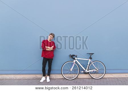 Attractive Young Man In A Red Sweatshirt Standing By A Bicycle On A Blue Background And Looking At T