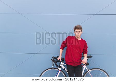 Portrait Of A Beautiful Guy In A Red Sweatshirt Standing With A White Bike On The Background Of A Bl