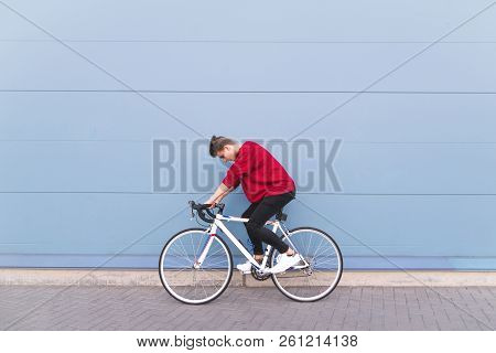 Young Man Riding A White Highway On A Background Of A Blue Wall. Stylish Hipster Cyclist Riding A Bi